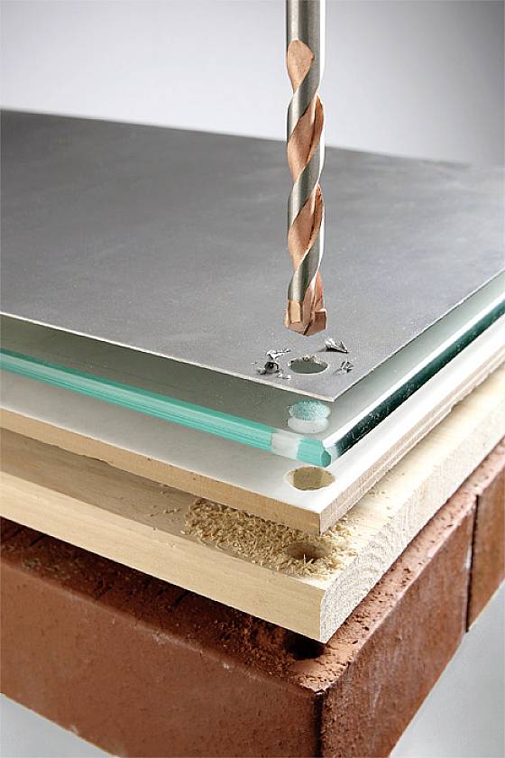 Bohrer durchdringt stapelweise Materialien: Metall, Glas, Holz und Ziegel. Demonstriert Vielseitigkeit und Stärke des Bohrers.