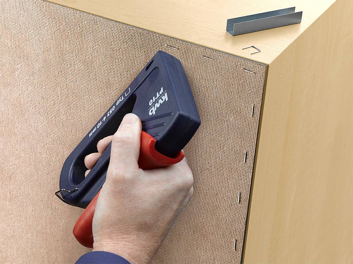 Eine Hand hält einen Tacker und befestigt Stoff mit Klammern an einer Holzkante. Oben liegen Ersatzklammern.