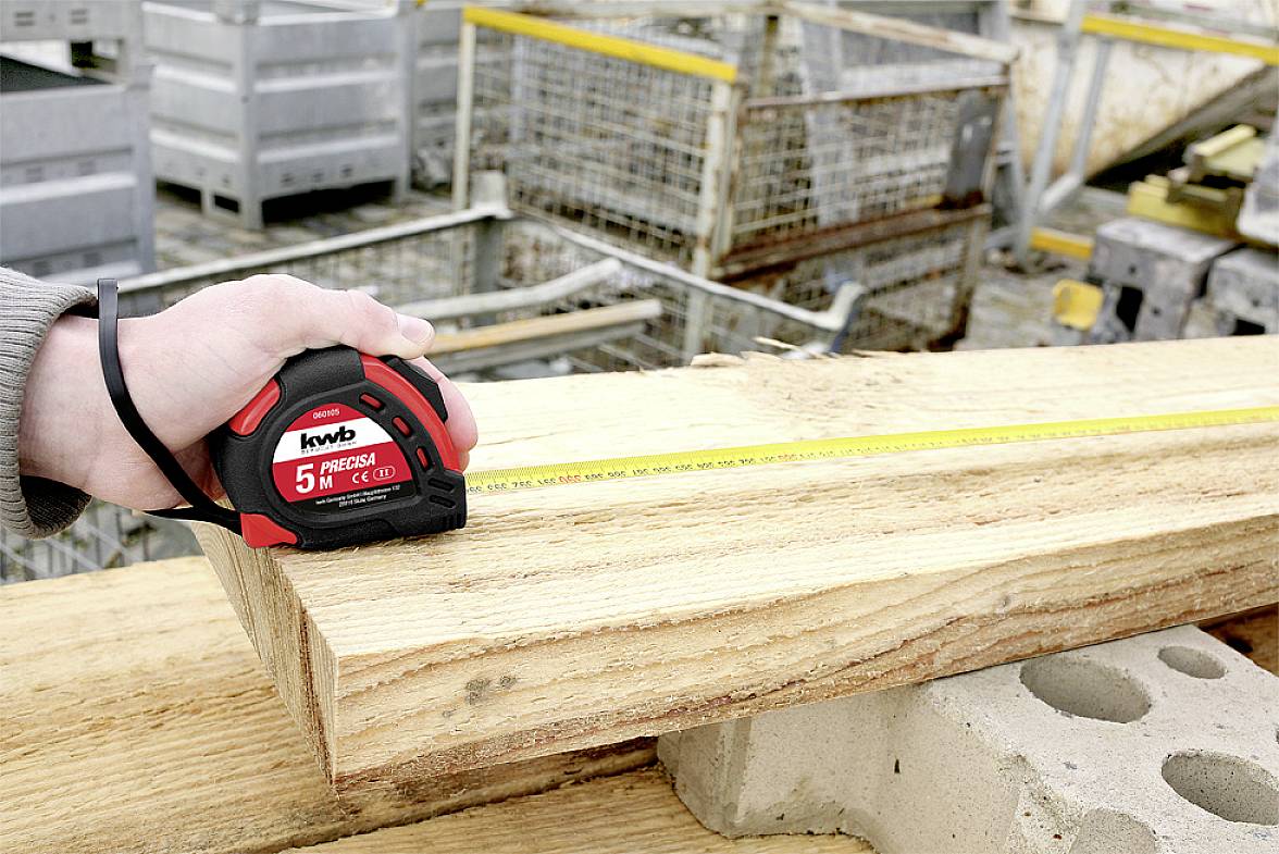 Eine Hand hält ein Maßband über ein Holzbrett und misst die Länge. Im Hintergrund sind Gitterboxen und weitere Bretter zu sehen.