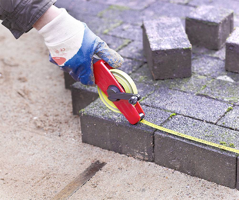 Eine Person vermisst mit einem Maßband Pflastersteine auf einem Gehweg. Die Pflastersteine sind dunkelgrau und der Boden ist sandig.