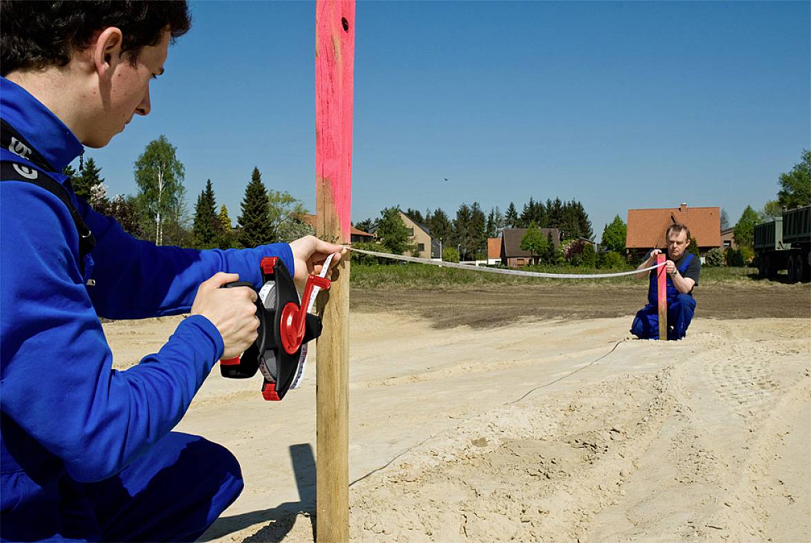 Zwei Personen in blauer Arbeitskleidung messen mit einem Bandmaß die Distanz zwischen einem Holzpfosten und einem Punkt am Boden auf einer Baustelle.