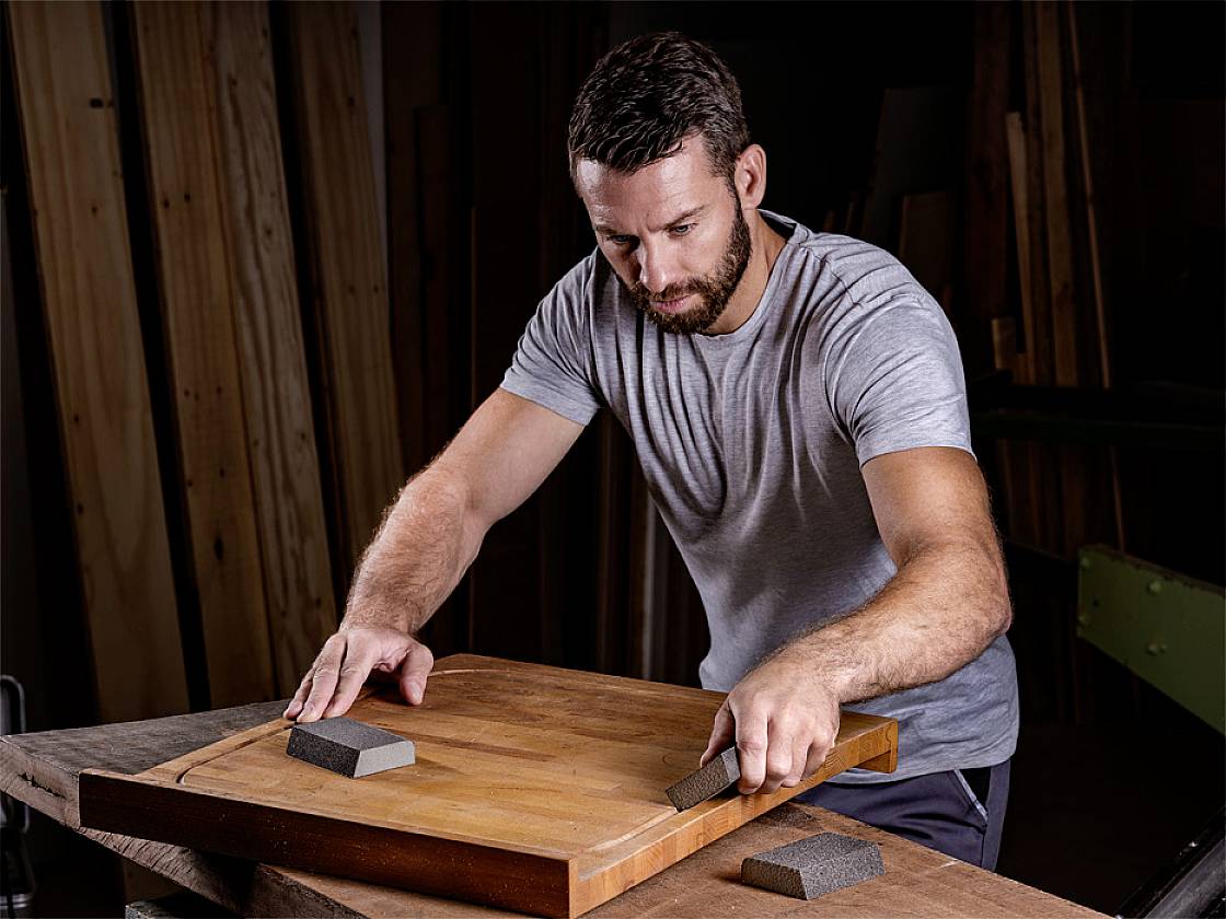 Ein Mann schleift mit Schleifblöcken ein Holzbrett in einer Werkstatt. Im Hintergrund sind Holzlatten zu sehen.