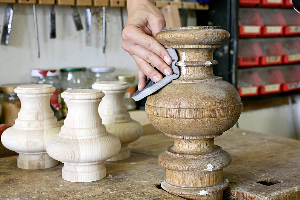 Eine Hand schleift ein großes, gedrechseltes Holzstück auf einer Werkbank. Im Hintergrund stehen weitere kleinere Holzteile.