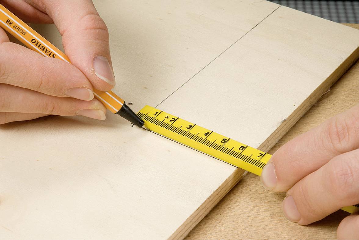 Eine Person markiert mit Bleistift und Maßband eine Linie auf einem Holzbrett zur Vorbereitung eines Schnitts.