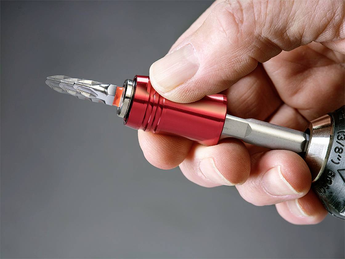 Eine Hand hält ein rot-metallisches Werkzeug mit einem Bohreraufsatz, bereit zur Verwendung. Im Hintergrund ist ein grauer unscharfer Bereich.