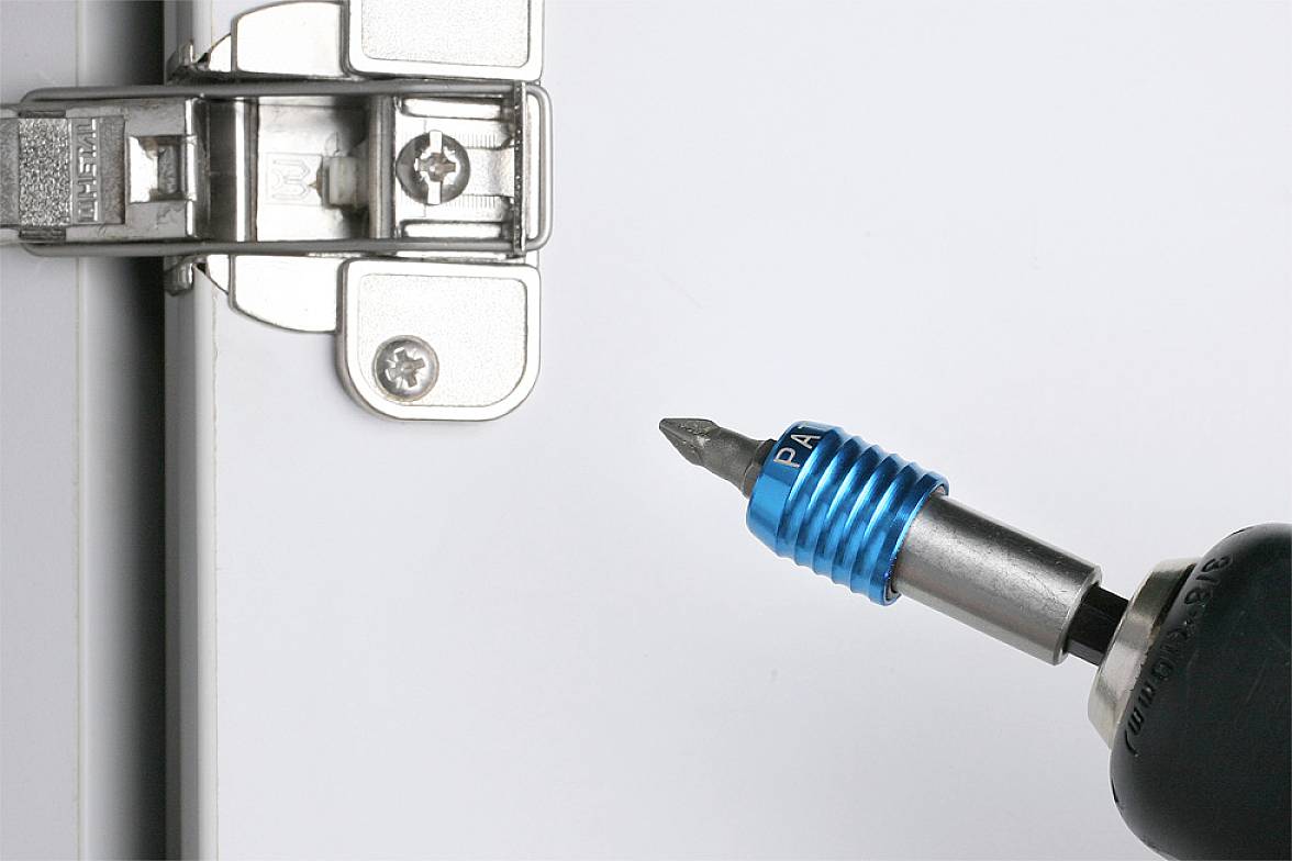 Ein blauer magnetischer Bit-Halter fixiert eine Schraube, während ein Akkuschrauber sie in ein Metallscharnier auf einer Tür einsetzt.