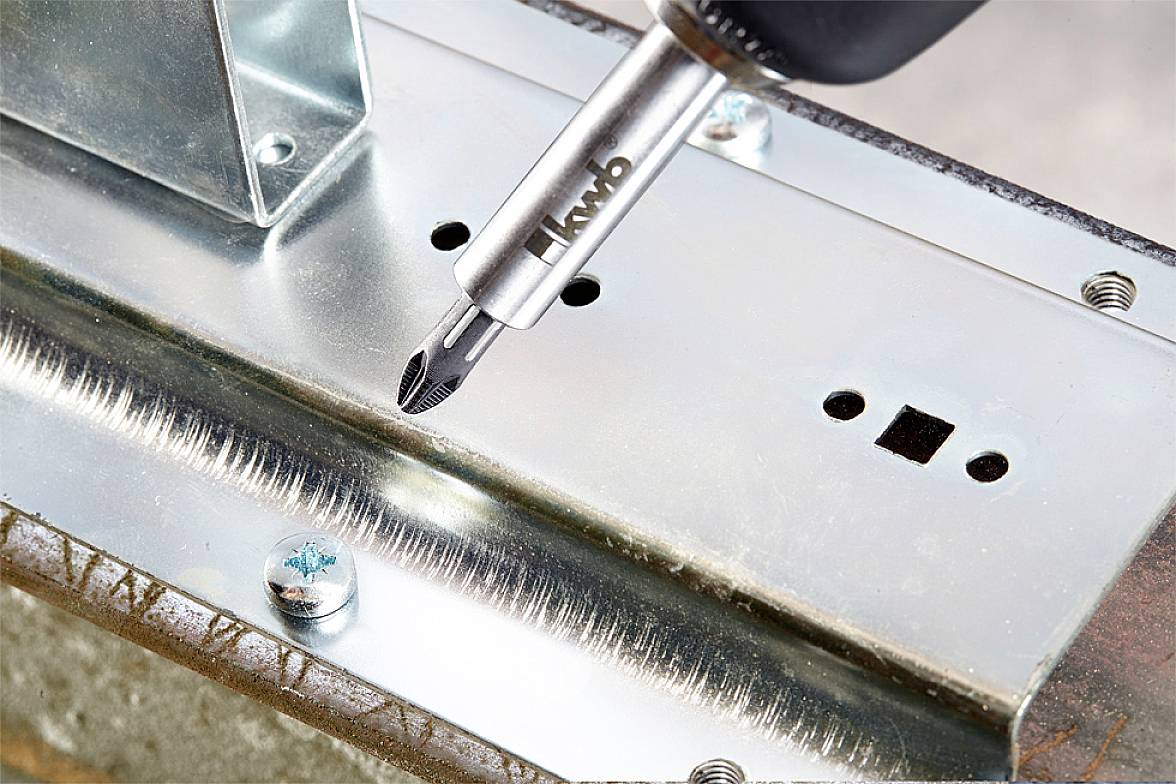 Eine Nahaufnahme eines Bohrers, der in einer Metallschiene positioniert ist. Der Bohrer trägt die Aufschrift 'kwb'. Schraube in der Nähe.