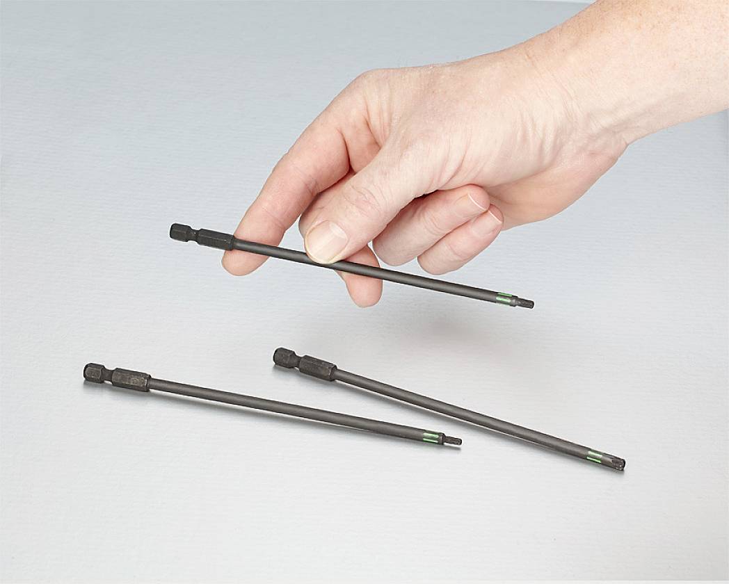 Eine Hand hält einen langen, dünnen Schraubendreherbit, während drei ähnliche Bits auf einer glatten Oberfläche liegen.