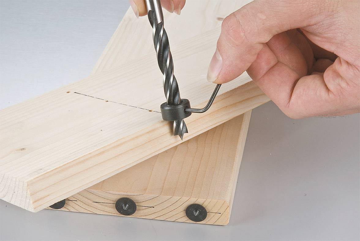 Eine Hand hält einen Bohrer, der ein Loch in zwei aufeinandergelegte Holzplatten bohrt. Ein Ring begrenzt die Tiefe des Bohrlochs.