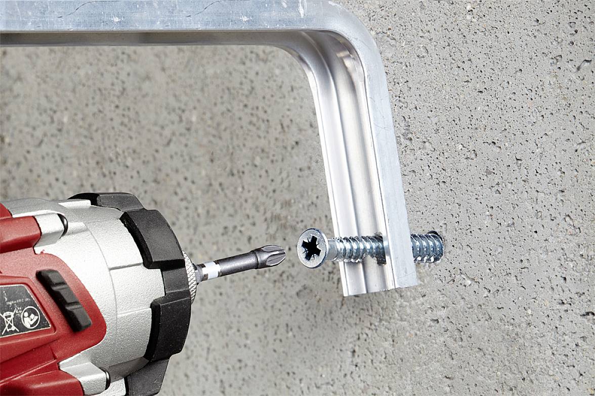 Ein Akkuschrauber zieht eine Schraube durch ein Metallwinkelstück in eine Betonwand. Der Bohrer dreht die Schraube ein.
