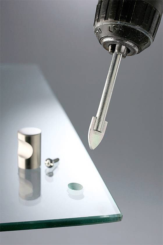 Ein Bohrer mit einem Glasbohrer bohrt ein Loch in eine Glasscheibe. Daneben liegen ein Glasverbinder und eine Schraube.