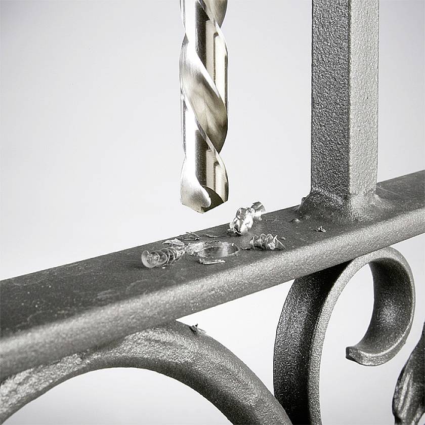 Ein Bohrer durchbohrt ein Metallgeländer, erzeugt Späne. Das Bild zeigt handwerkliche Präzision und die Struktur des Metalls.