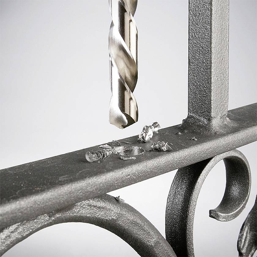 Ein Bohrer durchbohrt ein Metallgeländer, erzeugt Metallspäne; Fokus liegt auf dem Bohrvorgang und Spänen.