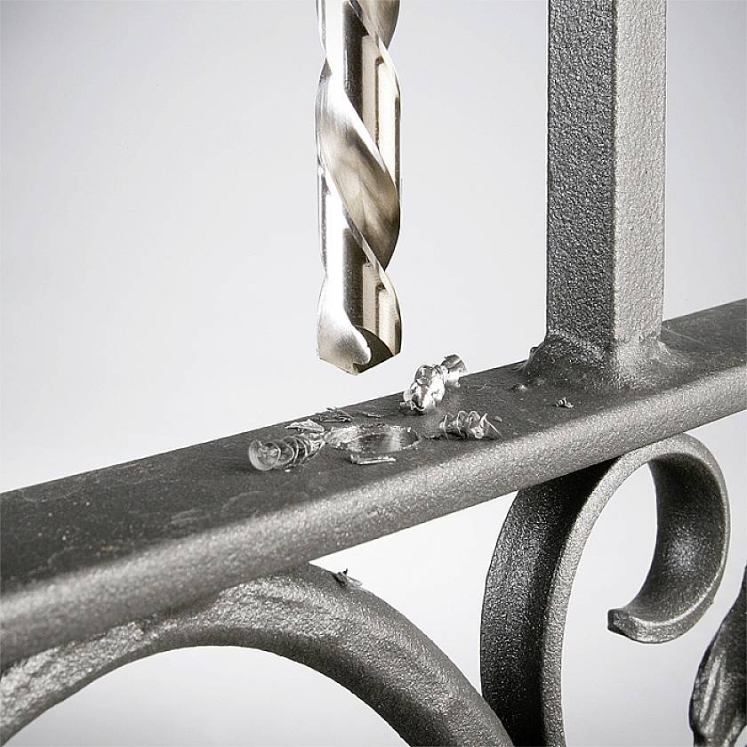 Ein Bohrer hinterlässt Metallspäne auf einer Metalloberfläche, die Teil eines dekorativen Geländers ist.