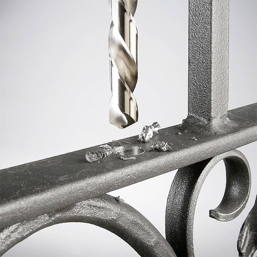 Ein Metallbohrer durchbohrt ein Stahlgeländer. Metallspäne sind um das Bohrloch verteilt.
