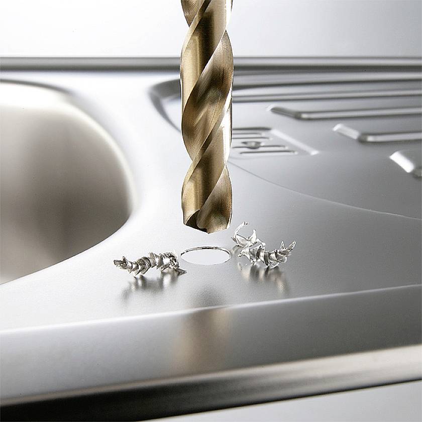 Ein Bohrer durchbohrt ein Edelstahlspülbecken. Metallspäne liegen um das Loch herum.