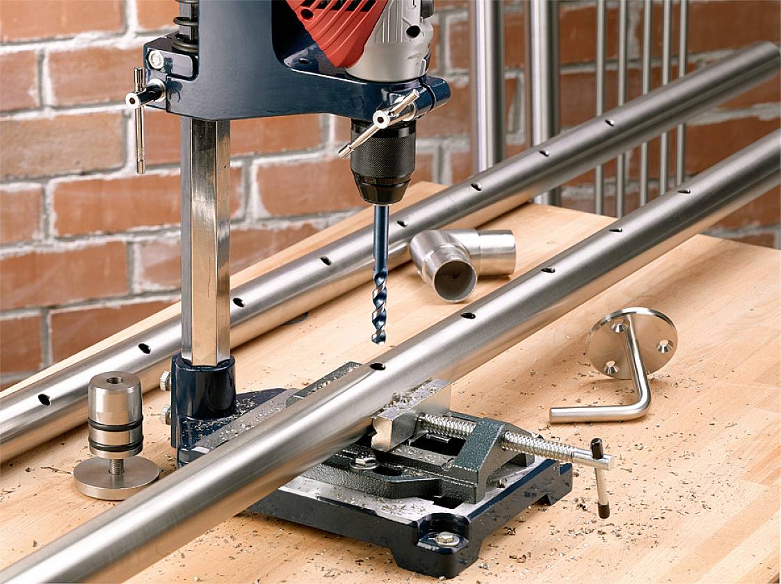 Metallbohrmaschine auf Werkbank, bohrt Löcher in ein Edelstahlrohr. Metallspäne um den Bohrer, Schraubstock hält das Rohr fest.