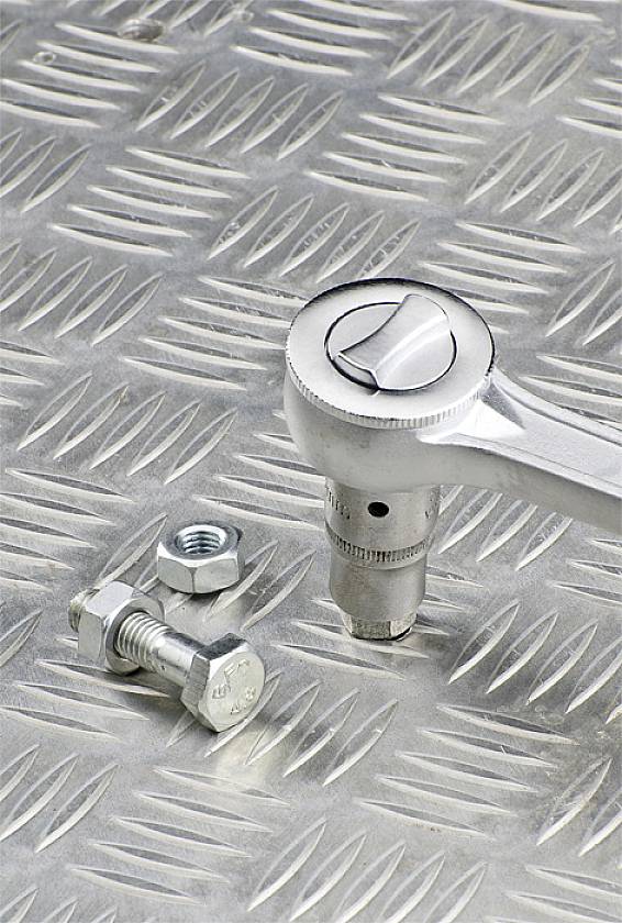 Eine Ratsche liegt auf einer Metallriffelplatte neben einer lose platzierten Schraube und Mutter.