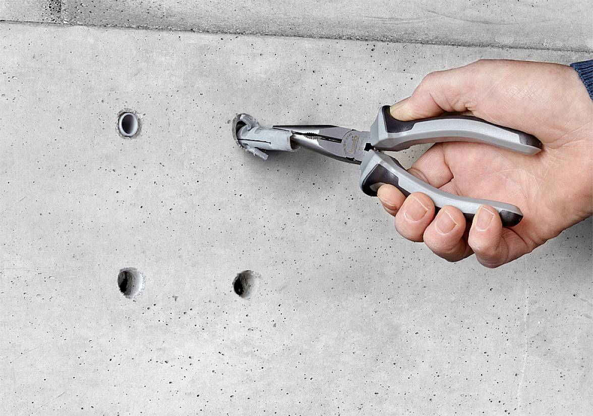 Eine Hand hält eine Zange und zieht einen Dübel aus einer Betonwand. Drei weitere Löcher sind in der Wand sichtbar.