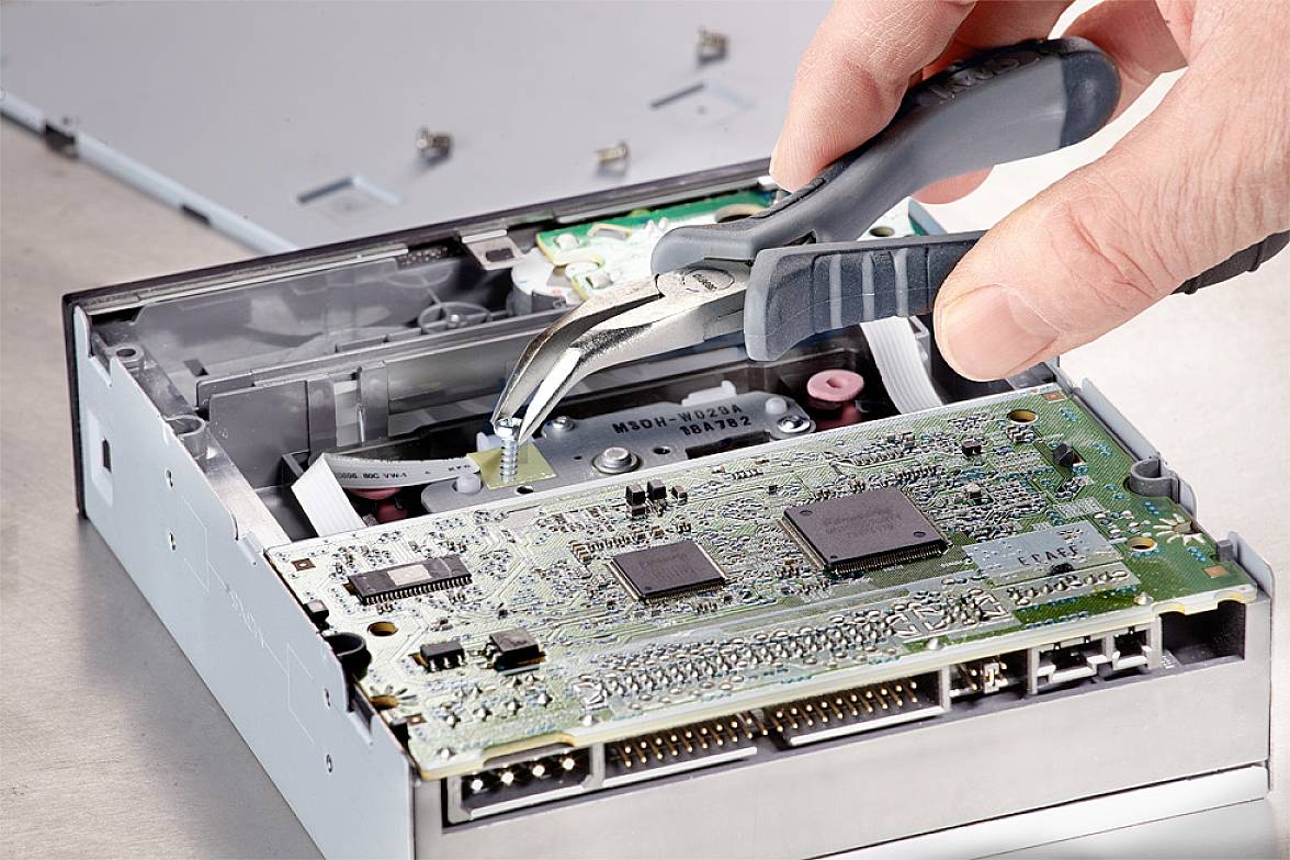 Eine Hand benutzt eine Zange, um an der offenen Elektronik eines Diskettenlaufwerks zu arbeiten, was zeigt, wie Hardware repariert wird.
