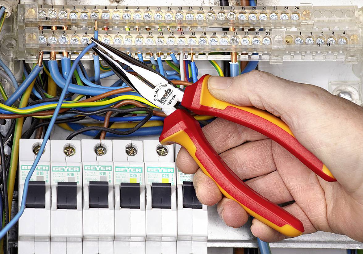 Eine Hand hält eine Spitzzange und arbeitet an Kabeln in einem Schaltschrank, umgeben von farbigen Drähten und Sicherungsautomaten.