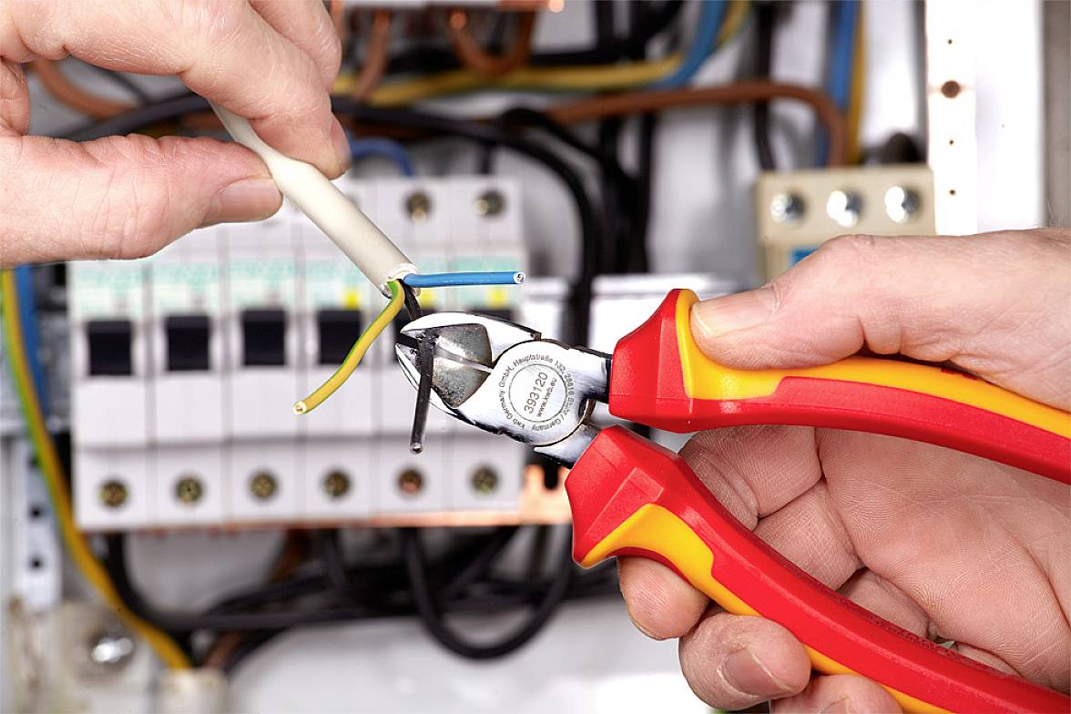 Zwei Hände halten einen Drahtschneider und schneiden ein Kabel vor einem elektrischen Sicherungskasten ab.