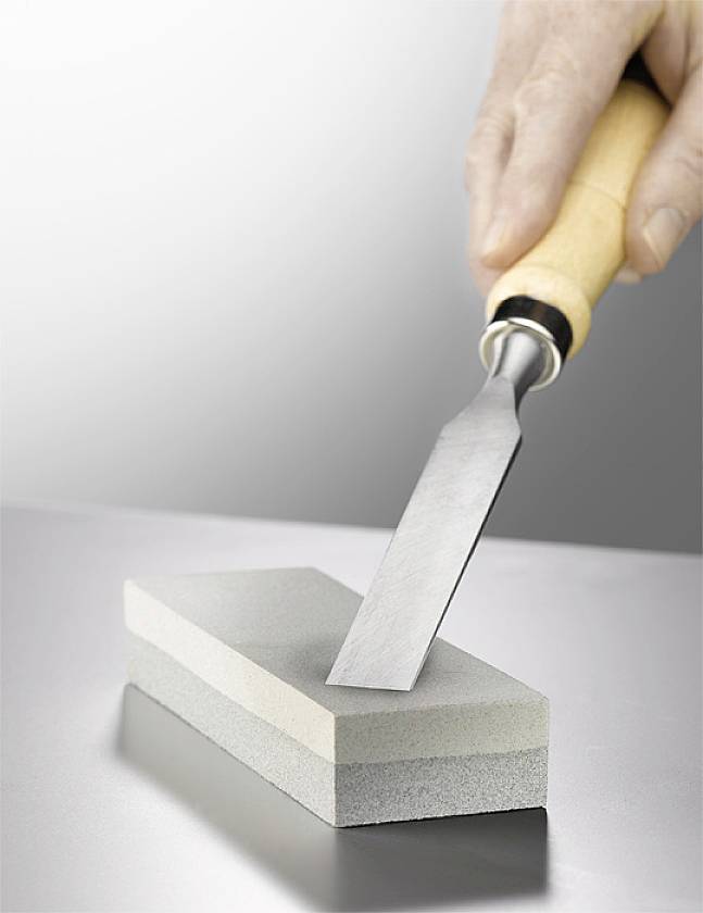 Eine Hand schärft ein Stechbeitel auf einem Schleifstein. Der Schleifstein liegt auf einer grauen Oberfläche.