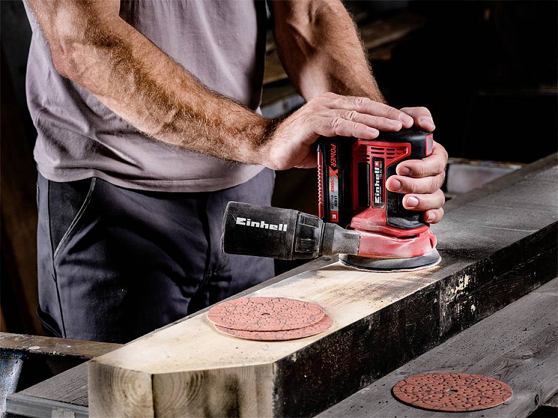 Eine Person schleift mit einem Einhell-Schleifgerät auf einem Holztisch. Schleifpapier und Holzstück sind ebenfalls zu sehen.