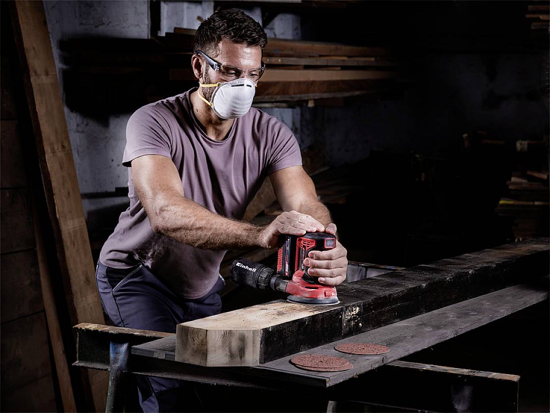 Ein Mann schleift ein Holzbrett mit einer elektrischen Schleifmaschine in einer Werkstatt. Er trägt eine Schutzbrille und eine Atemmaske.