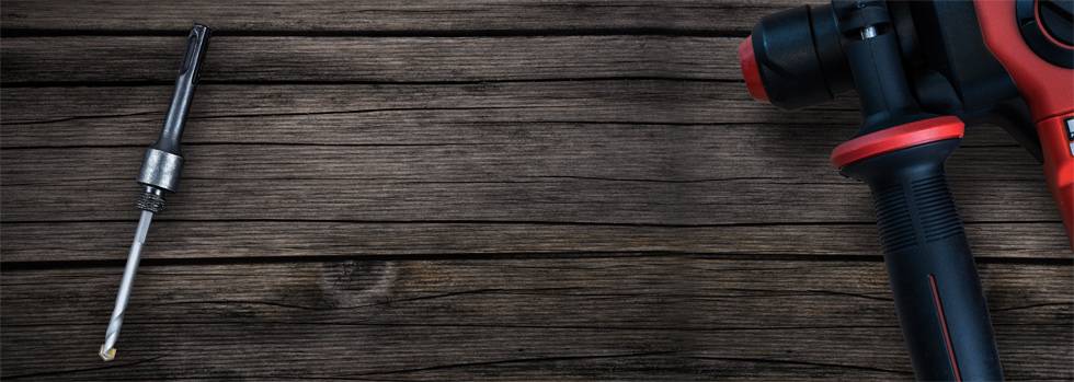 Eine rote Bohrmaschine mit schwarzem Griff liegt rechts auf Holzoberfläche, links daneben ein Bohreraufsatz.
