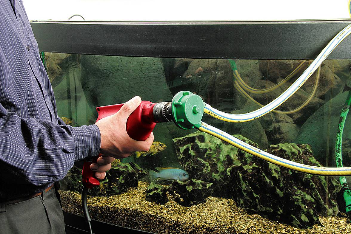 Eine Person hält ein rotes Werkzeug an einem Aquarium, während Schläuche ins Wasser führen. Ein Fisch schwimmt im Hintergrund.
