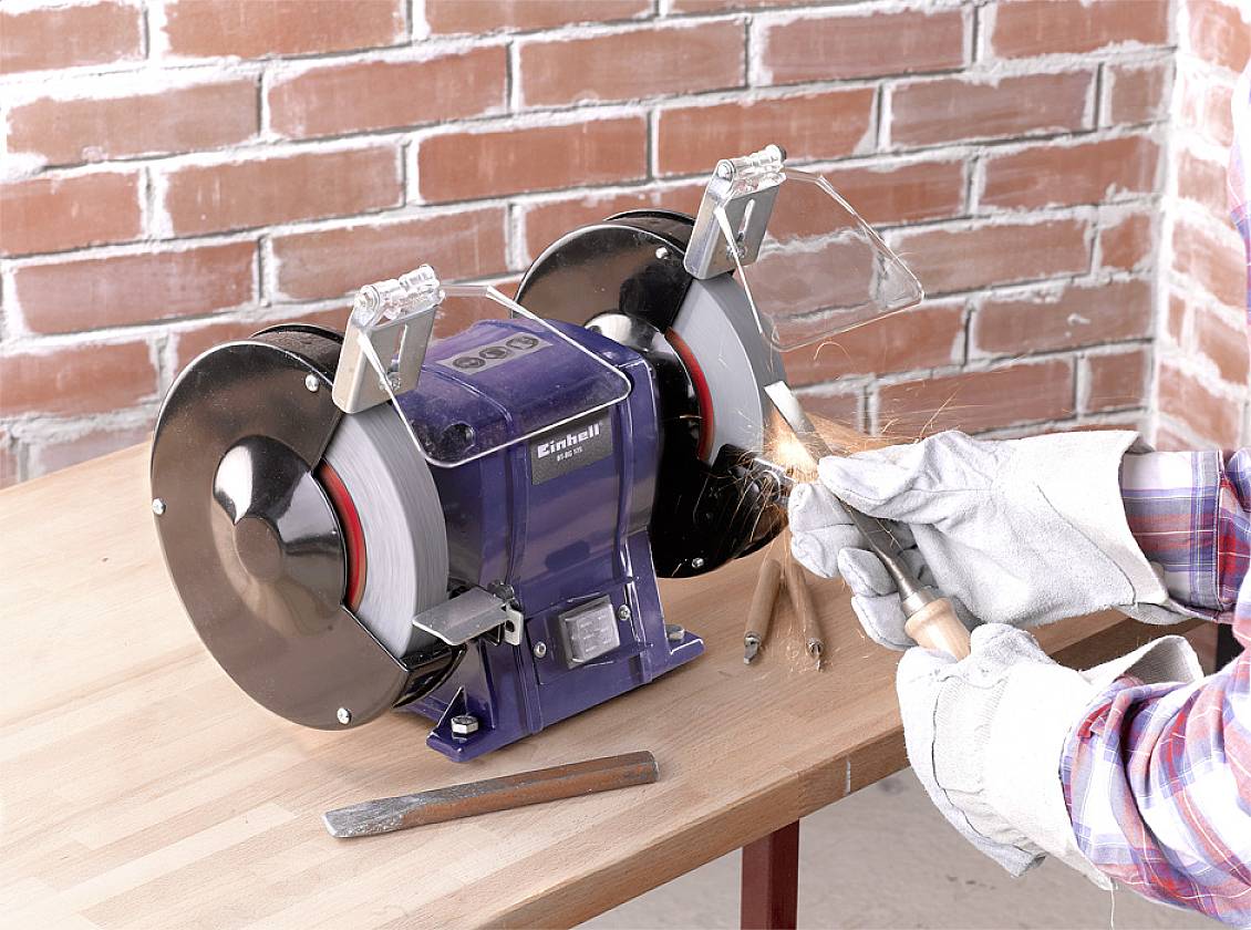 Eine Person schleift Metall auf einem doppelseitigen Schleifbock mit Funkenbildung. Der Schleifbock steht auf einem Holztisch.