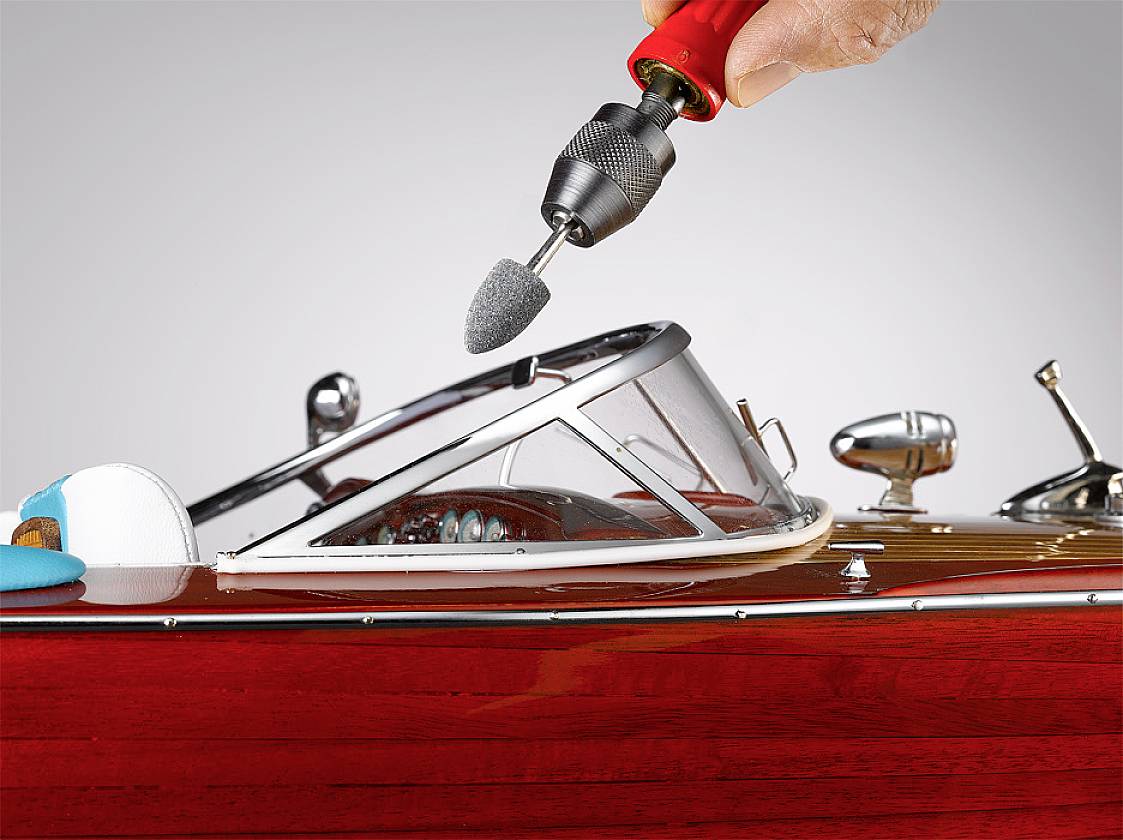 Ein Modellboot wird mit einem rotierenden Werkzeug bearbeitet. Eine Hand hält das Werkzeug, während der Fokus auf der präzisen Bearbeitung des Boots liegt.