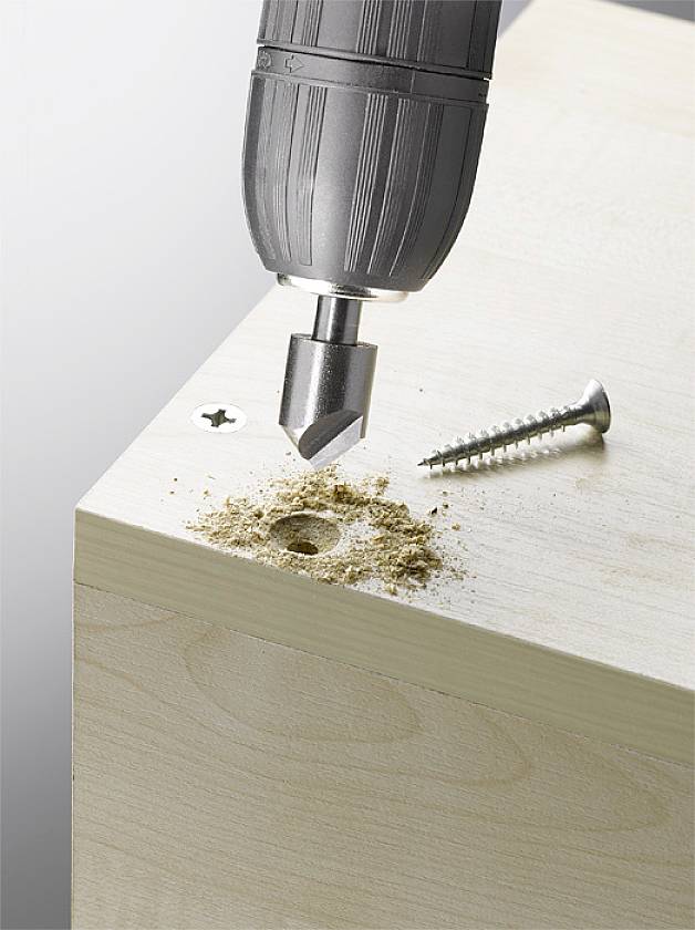 Ein Bohrer senkt eine Schraube in ein hölzernes Brett ein, daneben liegt eine lose Schraube. Holzspäne sind sichtbar.
