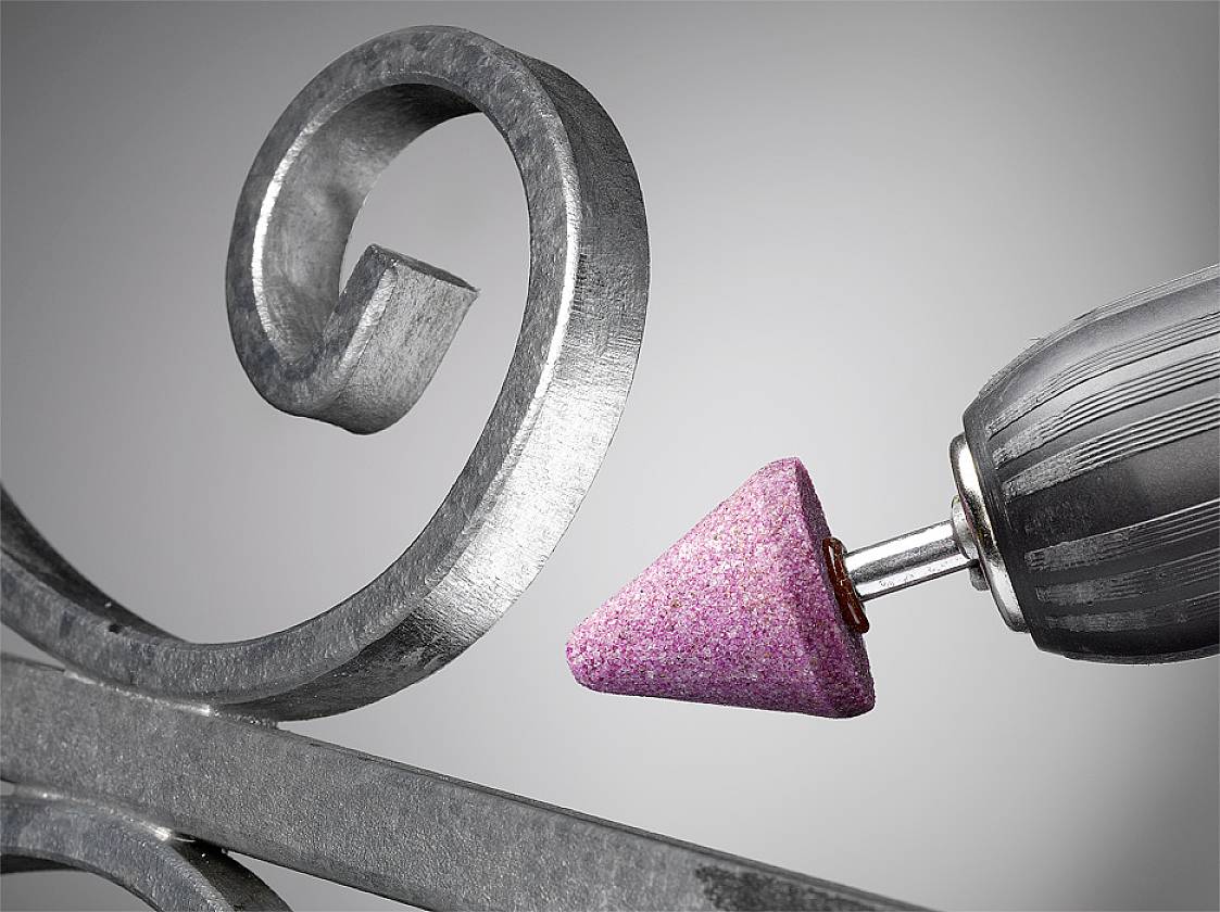 Ein elektrischer Schleifbohrer mit rosa Schleifkopf bearbeitet eine Metalloberfläche mit dekorativer Spirale.