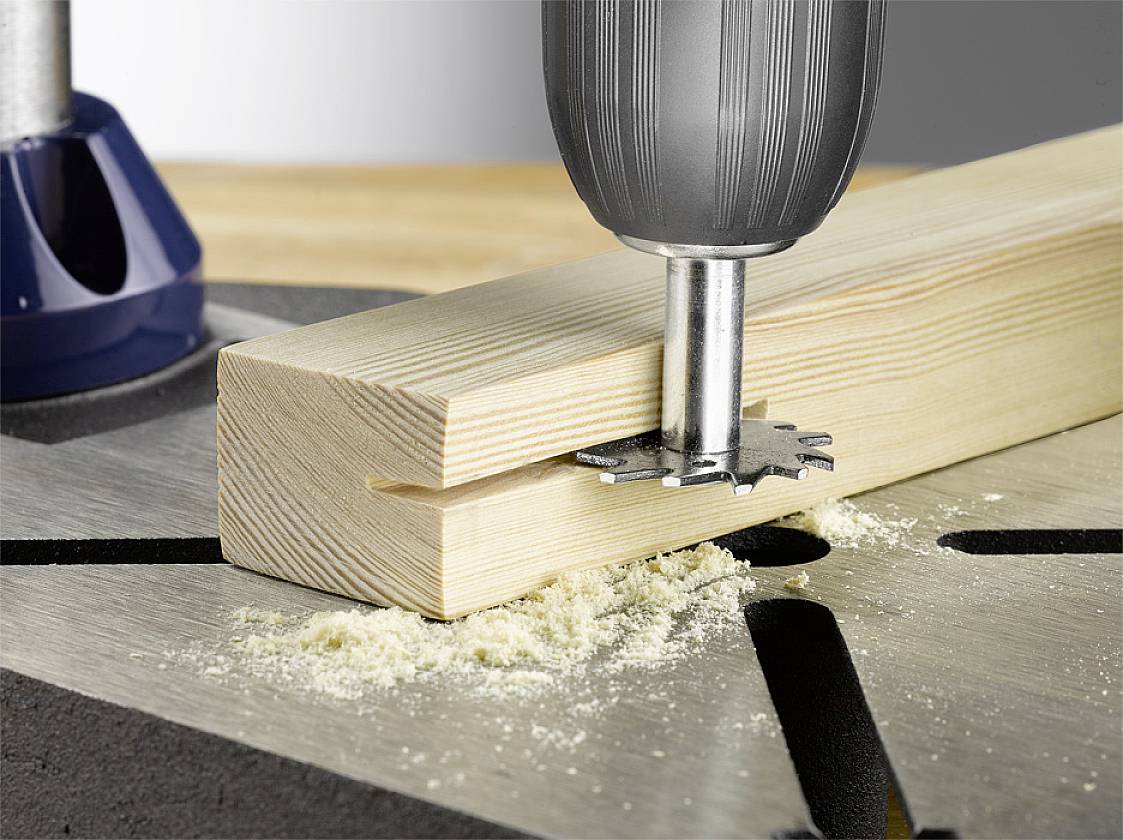 Ein Bohrkopf bearbeitet ein Holzstück, bohrt ein Loch. Holzspäne sind sichtbar.