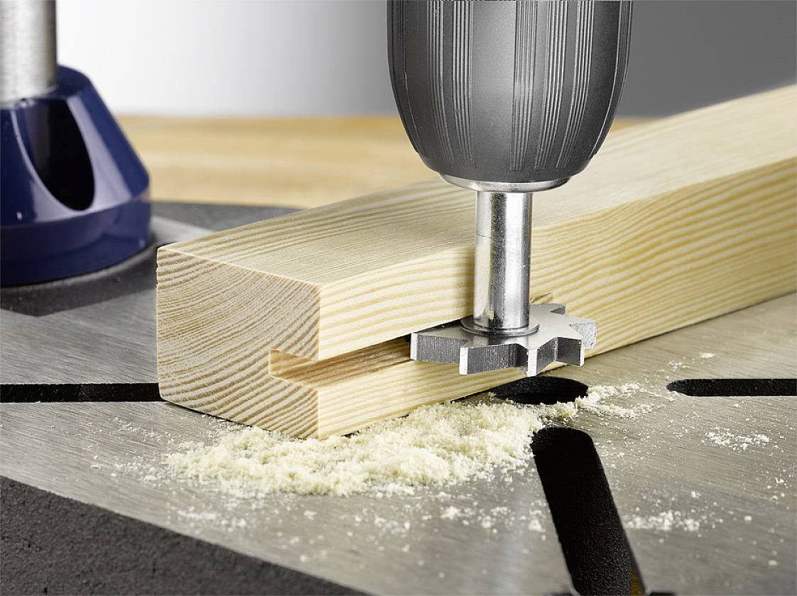 Ein Bohrer mit einem Fräsaufsatz arbeitet an einem rechteckigen Holzstück auf einer Werkbank, das Holz wird präzise bearbeitet.