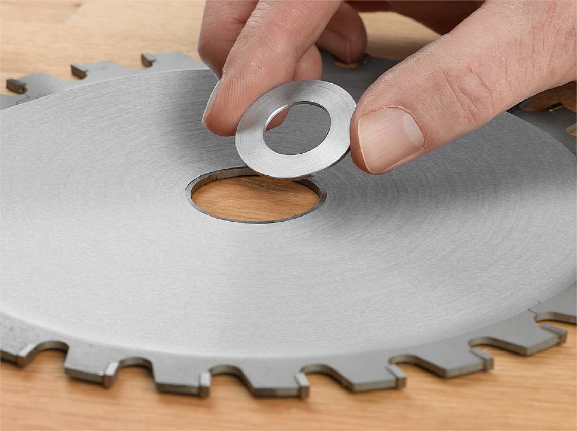 Eine Hand setzt einen kleinen Metallring auf das Loch einer größeren Sägeblatt-Scheibe, die auf einem Tisch liegt.