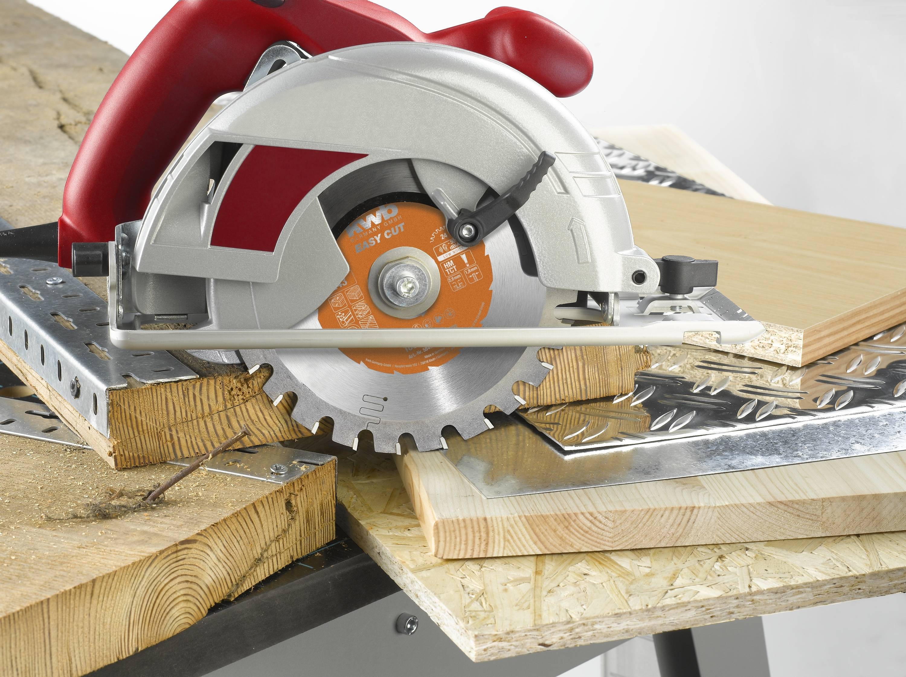Kreissäge auf Holztisch, sägt Holz und Metall. Roter Griff, silbernes Gehäuse. Umgebung ist Werkstatt mit verschiedenen Materialien.