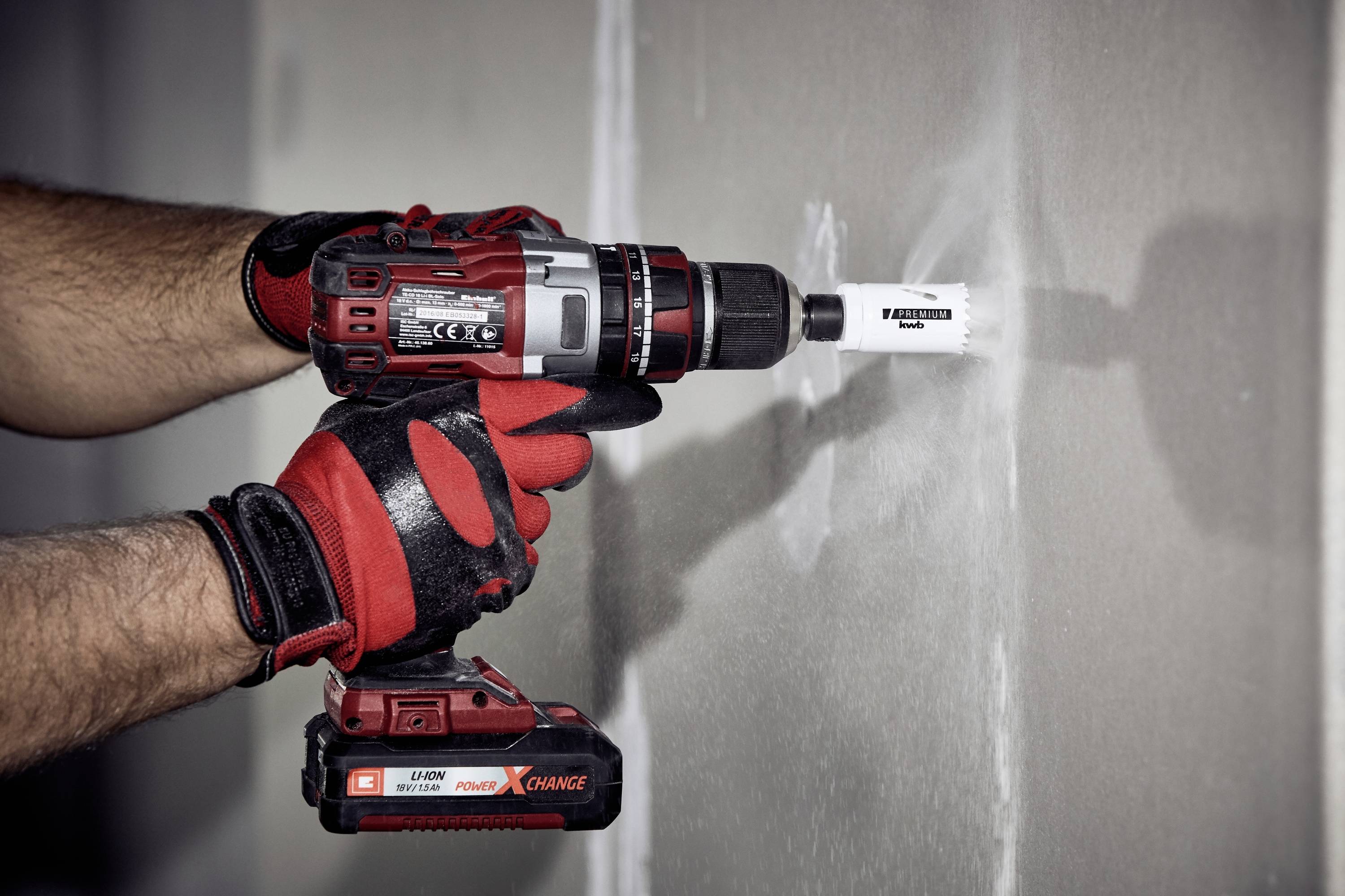 Ein Bohrer mit rotem und schwarzem Design wird von einer Person mit roten Handschuhen benutzt, um ein rundes Loch in eine Wand zu bohren.