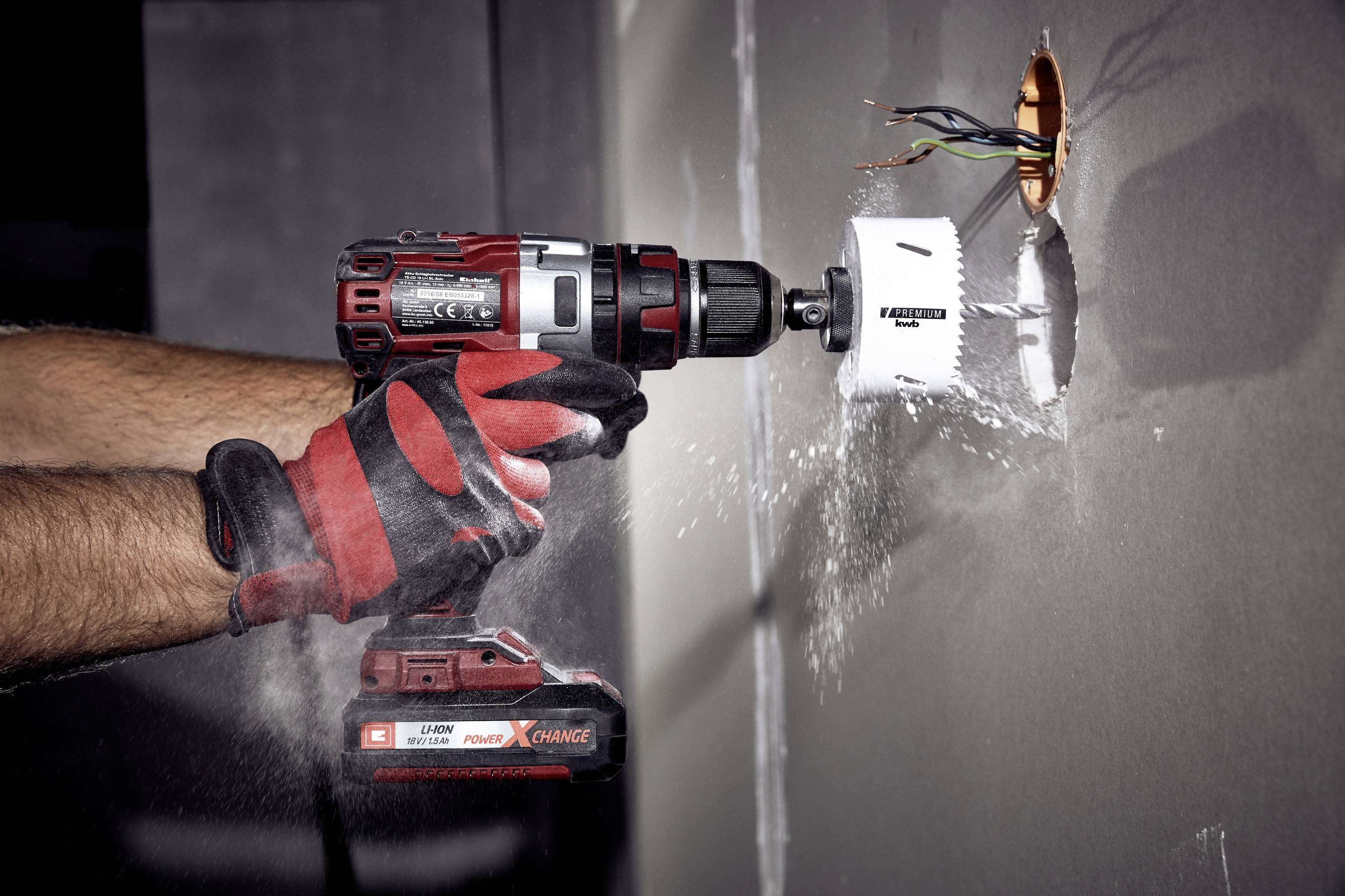 Eine Person bohrt mit einer Bohrmaschine ein rundes Loch in die Wand. Staub fliegt dabei auf. Kabel ragen aus der Wandöffnung.
