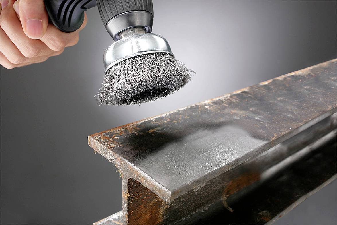 Eine Hand hält eine Drahtbürstenaufsatzmaschine und reinigt eine Metalloberfläche, um Rost zu entfernen. Glatte Fläche sichtbar.