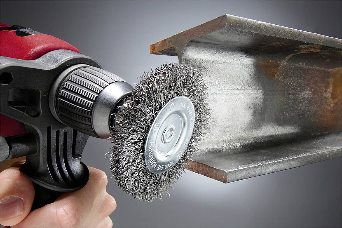 Eine Hand hält eine Bohrmaschine mit Drahtbürstenaufsatz, die Oberflächenrost von einem Metallträger entfernt.