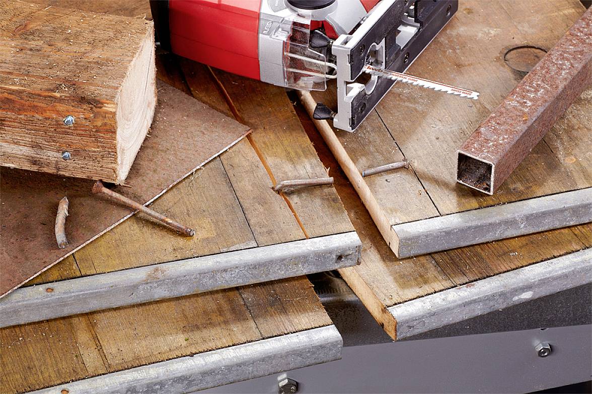 Bohrmaschine auf Holztisch, umgeben von Holzstücken und Nägeln. Ein Metallstück liegt daneben. Arbeitsplatz für Heimwerkerprojekte.