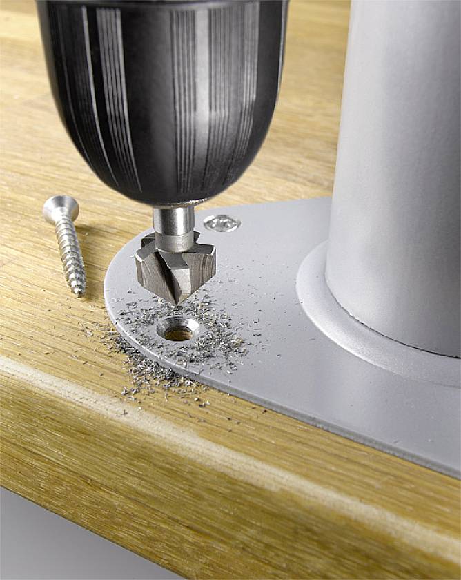 Eine Bohrmaschine senkt eine Schraube in eine Metallplatte ein, die auf Holz montiert ist. Daneben liegt eine weitere Schraube.