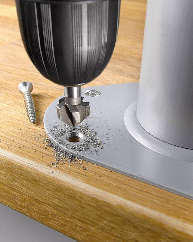 Bohrmaschine mit Senker fräst Loch an Metallkante; Holzuntergrund, Schraube daneben.