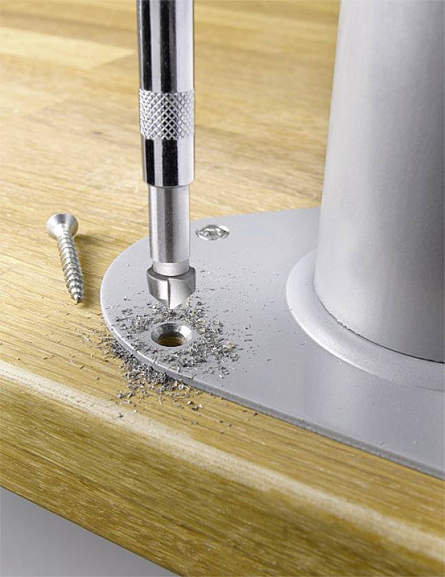 Ein Schraubenzieher dreht eine Schraube in ein Metallgestell auf einer Holzoberfläche, daneben liegen Metallspäne und eine weitere Schraube.