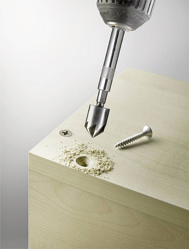 Ein Bohrer erzeugt ein Loch in einem Holztisch, daneben liegt eine Schraube.