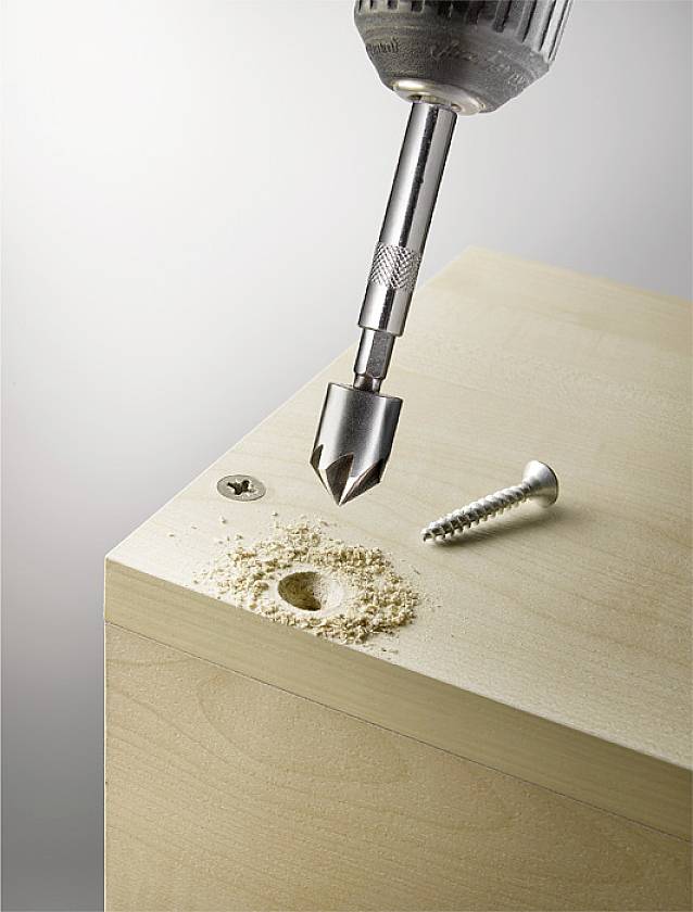 Ein Bohrer, der ein Loch in ein Holzbrett bohrt, mit Holzspänen daneben. Eine Schraube liegt bereit zum Eindrehen.