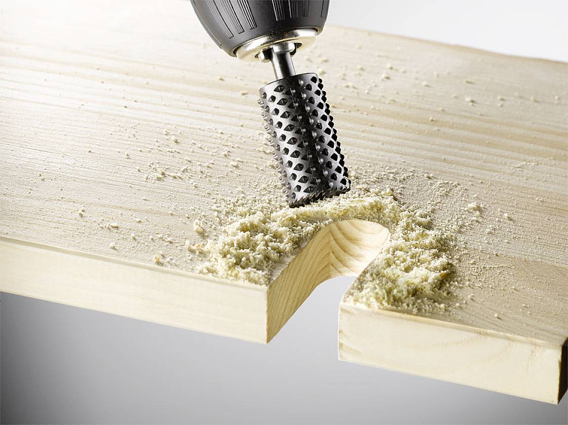 Ein Bohrer mit einer speziellen Fräse bearbeitet ein Holzbrett, wodurch Sägespäne entstehen. Die Fräse formt eine Mulde im Holz.